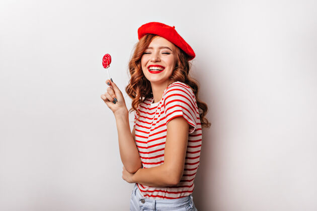 吃卷曲欢快的女孩用棒棒糖表达快乐可爱的法国年轻女子拿着糖果在白墙上笑甜点美味肖像