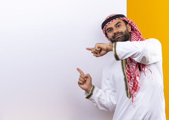 传统身着传统服装的阿拉伯商人站在空白广告牌旁 手指指着广告牌 脸上带着自信的微笑 越过橙色的墙壁脸指点商务人士