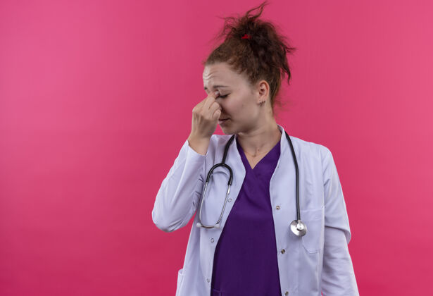 触摸年轻的女医生穿着白大褂 戴着听诊器 看上去很疲倦 工作过度 站在粉红色的墙上闭着眼睛摸鼻子听诊器医生外套
