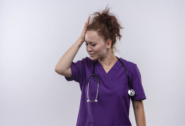 女人年轻的女医生穿着医疗制服 戴着听诊器 看起来很困惑 因为站在白墙上而摸了摸她的头触摸医生穿