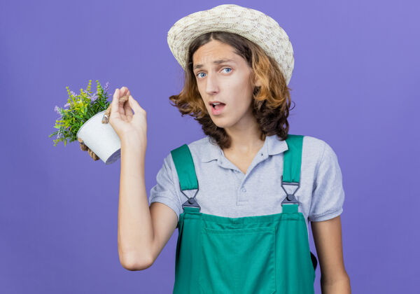 困惑年轻的园丁穿着连体衣戴着帽子拿着盆栽植物被搞糊涂了连身衣植物花园