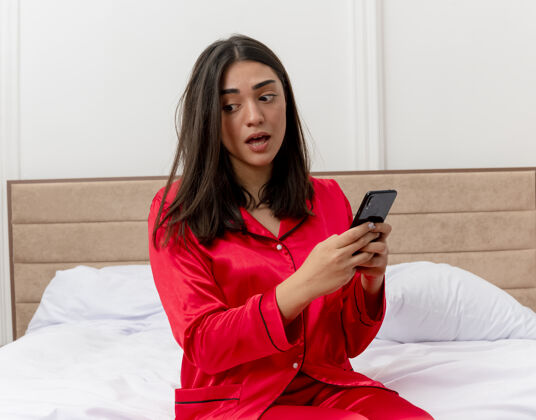 室内穿着红色睡衣的年轻美女坐在床上用智能手机看着困惑和非常焦虑的卧室室内灯光背景漂亮年轻女人