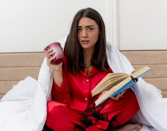 卧室穿着红色睡衣的年轻美女坐在床上 裹着毛毯 拿着一杯咖啡 看书 看着摄像机 在卧室的室内灯光背景下迷茫红色杯子毯子