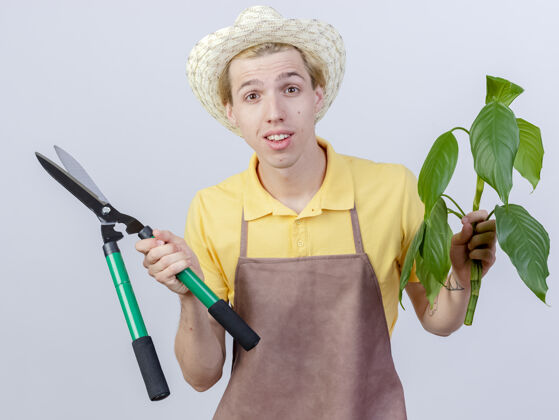 植物年轻的园丁身穿连体衣 戴着帽子 手持植物和树篱修剪器 站在白色背景上 面带微笑地看着镜头帽子篱笆快乐