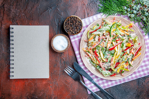 蔬菜顶视图蔬菜沙拉在盘子上桌布叉子和刀子盐和黑胡椒一个笔记本在深红色的桌子上食物菜肴盐