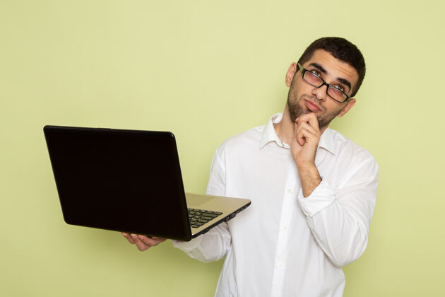 工作身穿白衬衫的男办公室工作人员手持并使用笔记本电脑在浅绿色墙上思考男性人笔记本电脑