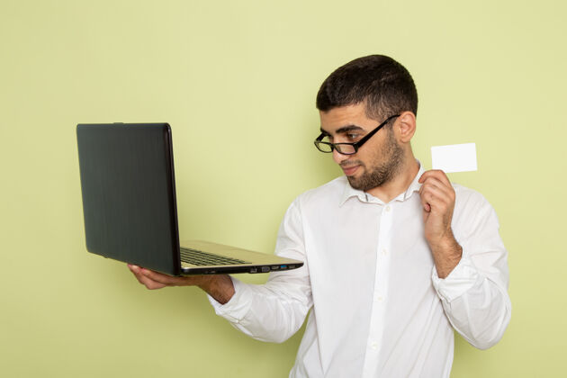 工作身穿白衬衫的男办公室工作人员手持笔记本电脑和卡片站在浅绿色墙上的正视图男性男士浅绿色