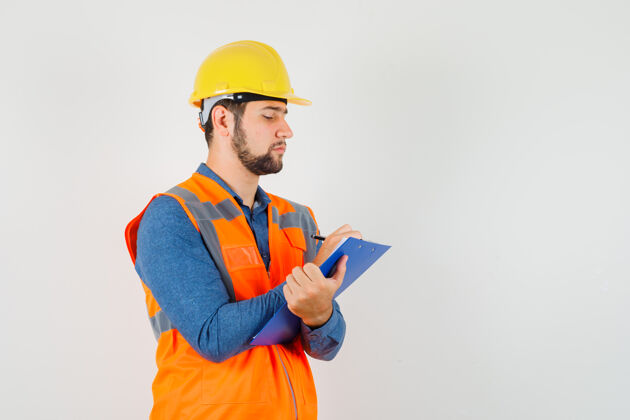 年轻人年轻的建筑工人穿着衬衫 背心 头盔 在剪贴板上做笔记 看起来很忙 正面视图安全工具努力