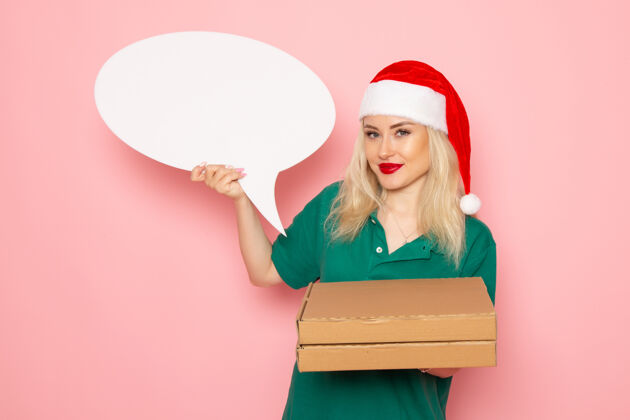 工作正面图年轻女性手持白色大牌子和食品盒在粉色墙上拍照工作制服新年假期工作快递盒子信使年轻的女性