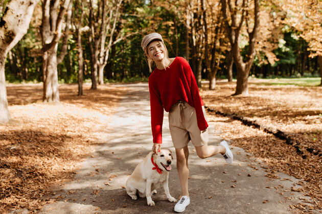 季节穿着时髦的红色毛衣和米色短裤的漂亮年轻女子和她的狗在美丽的秋天公园里玩得很开心公园腰漂亮