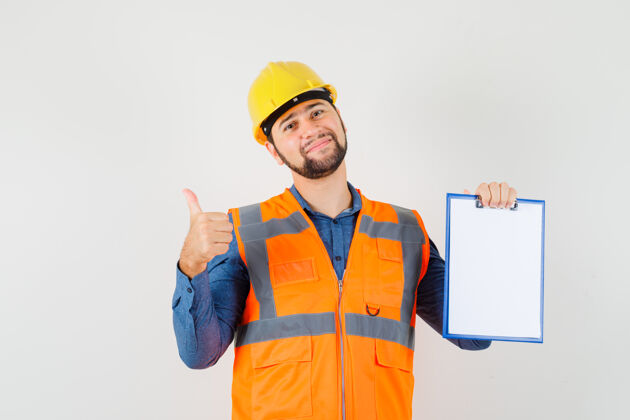 工厂年轻的建设者竖起大拇指 穿着衬衫 背心 戴着头盔拿着剪贴板 看上去很快乐 正前方的景色工人工厂工业