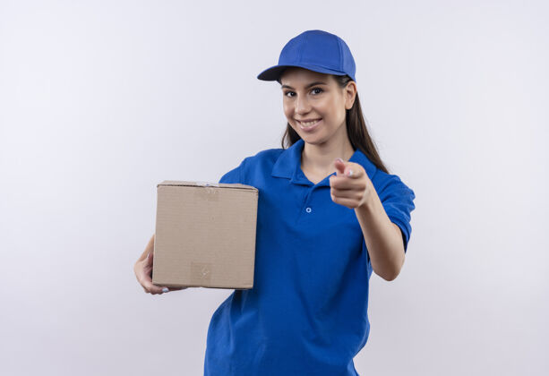 微笑身穿蓝色制服 戴着帽子的年轻送货女孩拿着盒子 微笑着自信地用手指着相机女孩站着制服