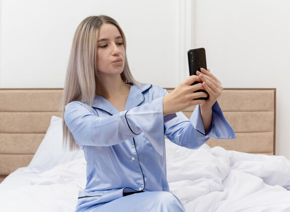 漂亮穿着蓝色睡衣的年轻美女坐在床上用智能手机自拍 在卧室内部灯光背景下吹一吻床卧室蓝色