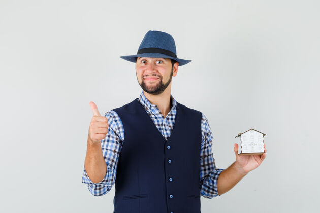 前面年轻人举着房子的模型 在衬衫 背心 帽子和看起来快乐 正面视图拇指向上男人模特休闲