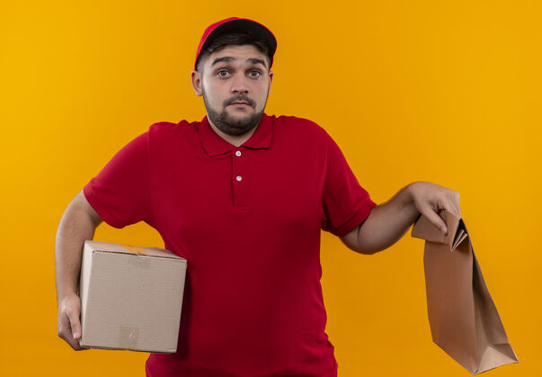 红色年轻的送货员穿着红色制服 戴着帽子 手里拿着盒子和纸袋 看上去既困惑又不确定盒子困惑年轻