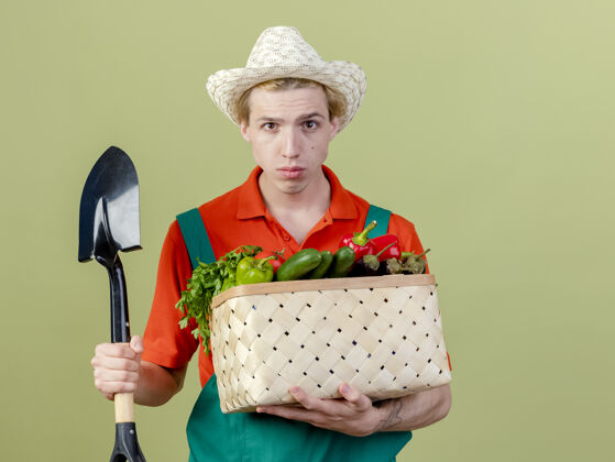 穿年轻的园丁穿着连体衣 戴着帽子 手里拿着装满蔬菜的箱子和铲子 站在灯光下严肃地看着摄像机看年轻人花园