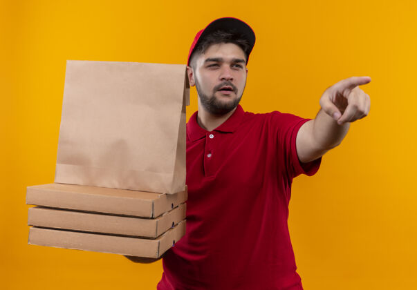 怀疑年轻的送货员穿着红色制服 戴着帽子 手里拿着纸包和一叠比萨饼盒 指着一个表情怀疑的东西纸什么指着