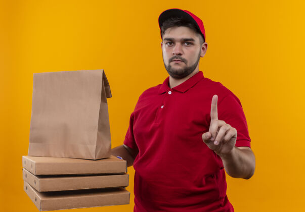 盒子年轻的送货员穿着红色制服 戴着帽子 手里拿着纸包和一叠披萨盒 表情严肃地用食指警告警告红色目录