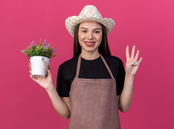 花盆面带微笑的白人女园丁 戴着园艺帽 拿着花盆 四个手指在粉色的墙壁上做手势 还有复制空间帽子四园艺