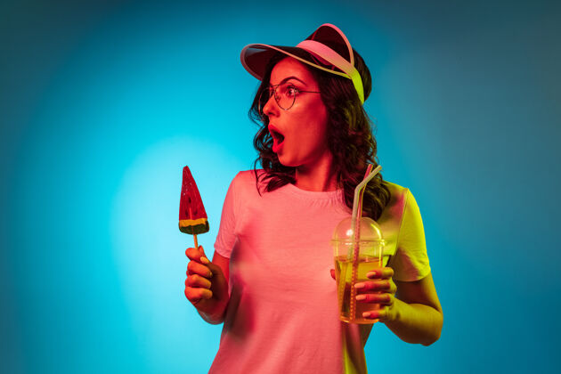 美女戴着帽子拿着糖果和饮料在时髦的蓝色霓虹灯工作室的年轻女子霓虹灯站成功