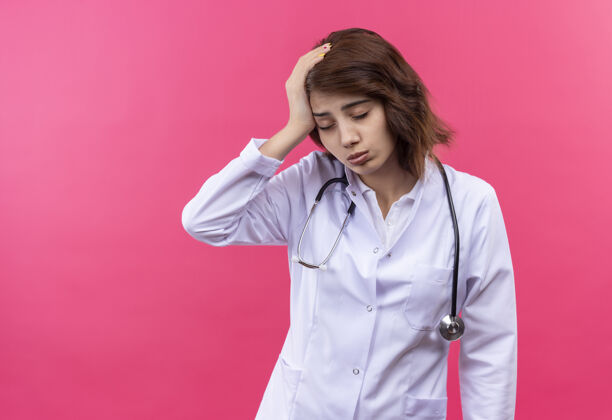 站着穿着白大褂 戴着听诊器的年轻女医生看起来很累 手放在头上 眼睛闭着 工作过度看外套医生