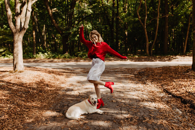 风穿着红毛衣的美丽幸福的金发女郎在公园里和她的拉布拉多犬一起享受美好时光穿着暖和衣服的时髦女孩在落叶间摆姿势毛衣季节欢乐