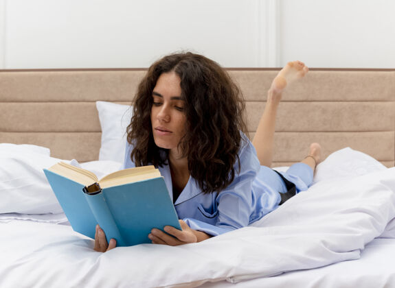 躺着穿着蓝色睡衣的年轻美女躺在床上 在卧室的室内灯光背景下看书睡衣女人蓝色
