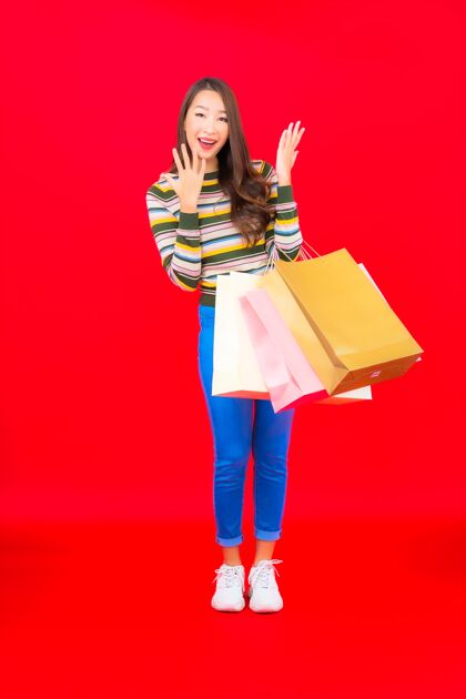 年轻在红墙上画一幅美丽的亚洲年轻女子 带着五颜六色的购物袋时尚购买购物狂