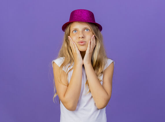 微笑戴着节日礼帽的漂亮小女孩抚摸着脸颊感受着积极的情绪微笑着 生日派对的概念站在紫色的背景上女孩生日节日