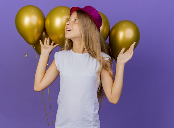 帽子戴着节日礼帽的漂亮小女孩和一大群巴隆人面带喜色 站在紫色背景下的生日派对概念漂亮生日看起来