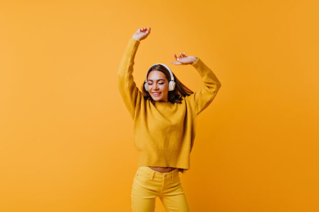 快乐可爱的黑发女士穿着黄色的裤子欣赏好歌穿着柔软毛衣的无忧无虑的女人在橙色的墙上跳舞服装放松时尚