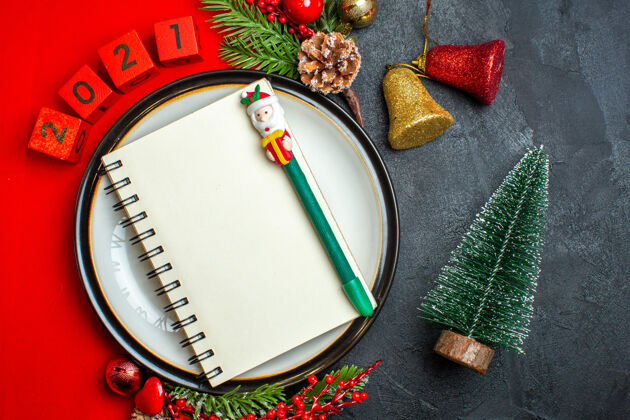 数字新年背景俯视图 笔记本上有钢笔 餐盘上有装饰配件 杉木树枝和数字放在黑色桌子上圣诞树旁的红色餐巾上手镯杉木视图