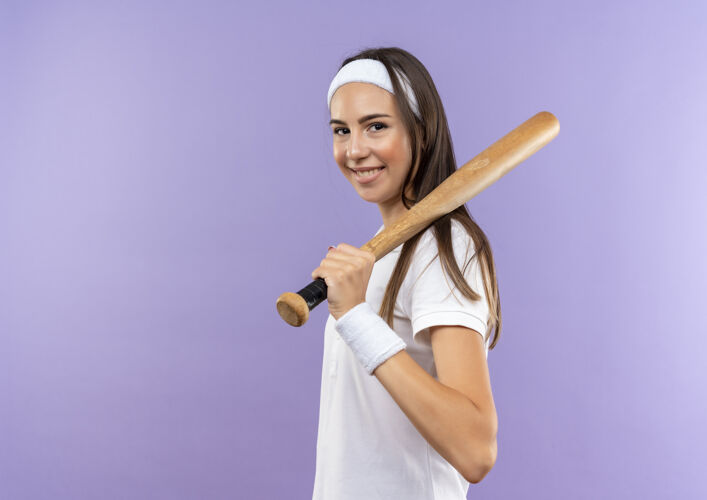 女孩微笑着漂亮的运动女孩戴着头带和手镯肩上拿着棒球棒站在侧面视图孤立的紫色空间头带腕带视图