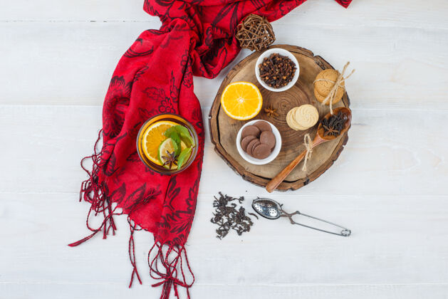 草药顶视图上有一碗饼干和丁香 木板上有柑橘类水果和凉茶 白色表面有红领巾和滤茶器健康装饰柑橘
