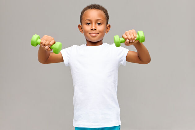 运动可爱的肖像适合运动深色皮肤的男孩在白色空白t恤做早晨锻炼常规的二头肌使用两个绿色哑铃有活力快乐的面部表情健身小学生孩子