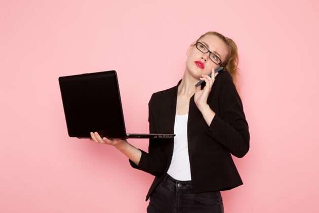 商务身穿黑色夹克的女上班族在粉色墙上使用笔记本电脑的正视图工作麦克风模特