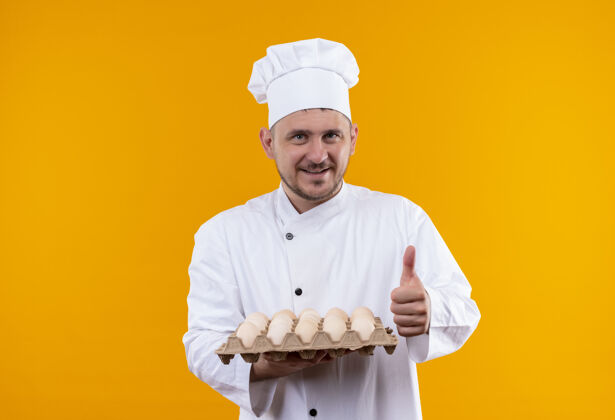 拇指身着厨师制服的年轻帅哥面带微笑 手里拿着一盒鸡蛋 在橙色的空间里孤立地竖起大拇指向上鸡蛋制服
