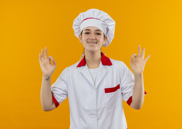 橙色微笑着的年轻漂亮厨师身着厨师制服 戴着牙套 在橙色的空间里做着“ok”的手势牙科好厨师