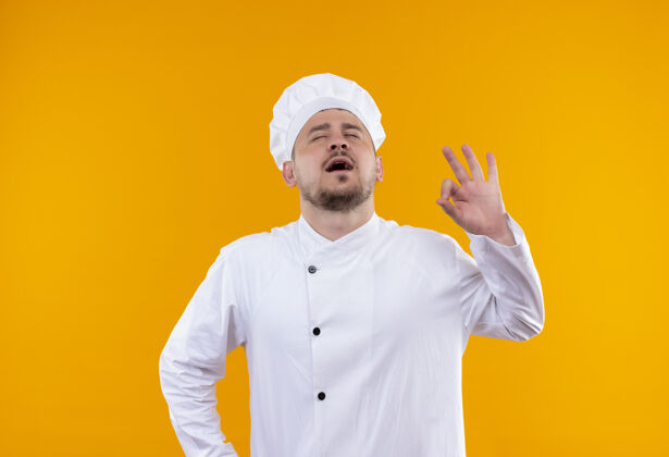 厨师穿着厨师制服的年轻帅气的厨师在孤立的橙色空间闭着眼睛张嘴做着“ok”的手势制服帅气厨师