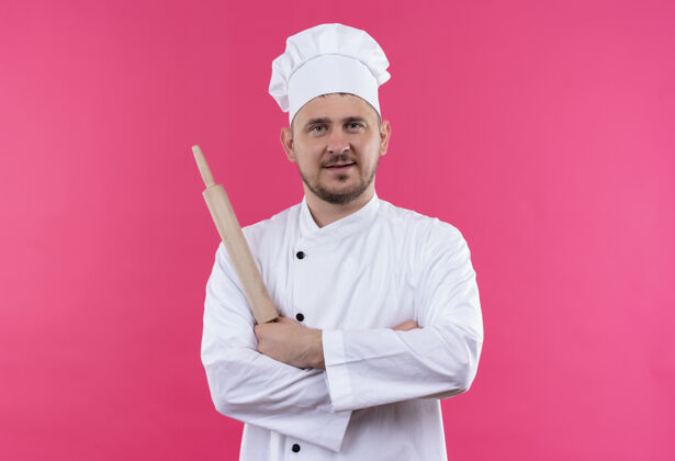 别针年轻帅气的厨师身着厨师制服 手持擀面杖 以封闭的姿态站在粉红色的空间里抱着年轻粉色