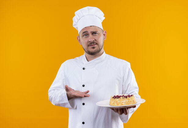 年轻年轻帅气的厨师穿着厨师制服 拿着蛋糕盘指着孤立在橙色空间厨师拿着蛋糕