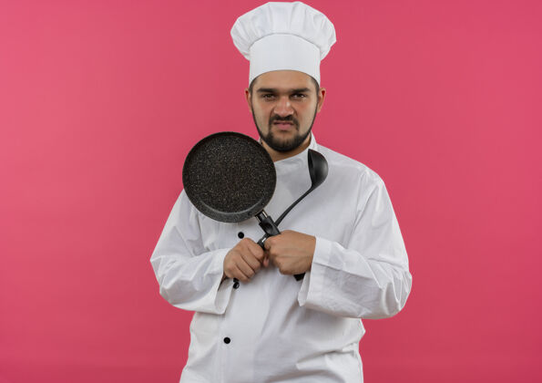 不愉快穿着厨师制服的年轻男厨师手拿煎锅和勺子 看上去在粉红色的空间里与世隔绝男性制服看