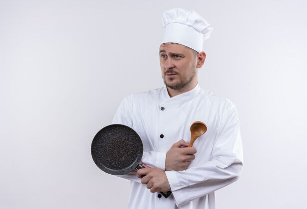 勺子年轻帅气的厨师穿着厨师制服 拿着煎锅和勺子 看着隔离在空白处的一边煎侧厨师