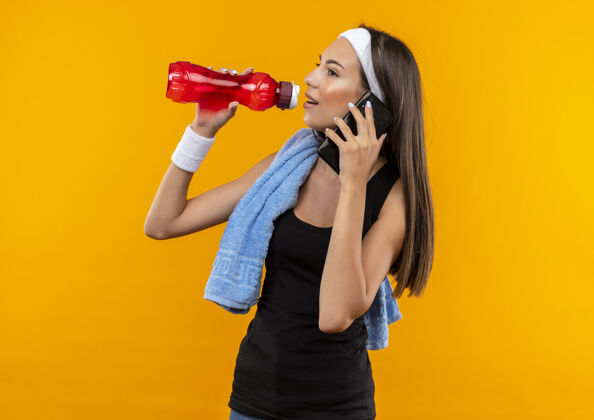 水高兴的年轻漂亮的运动女孩戴着头带和腕带试图喝瓶子里的水 并在电话里看着橙色空间孤立的一面饮料说话漂亮
