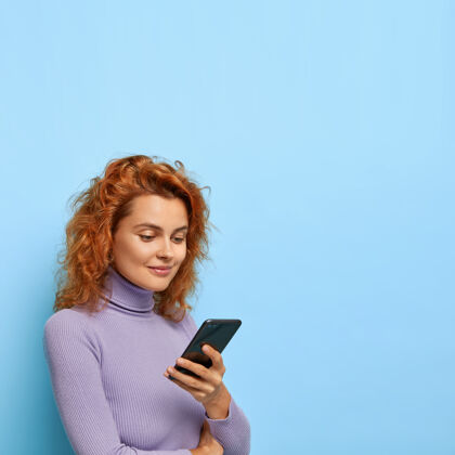 女孩可爱的姜女照片站在半转身 使用现代智能手机 检查邮箱 穿着休闲服 隔离在蓝色墙上 广告复制空间模特风格设备