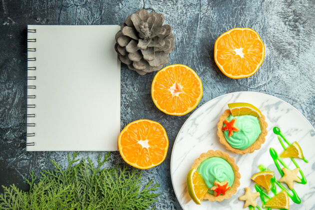 笔记本俯视图绿色糕点奶油小馅饼和柠檬片在盘子上切橘子笔记本在黑暗的桌子上糕点小果汁