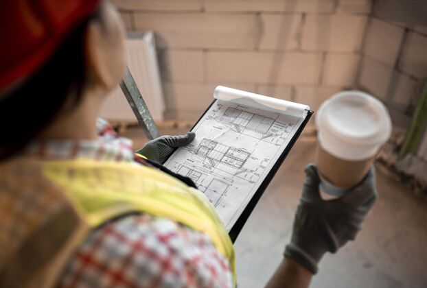 背心高角度的女建筑工人拿着咖啡和蓝图硬工作职业