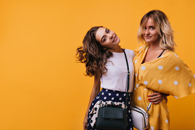金发感兴趣的黑发女人拿着时髦的手提包站在黄色的朋友旁边穿着橙色裙子的漂亮白种女人时尚兴奋女性