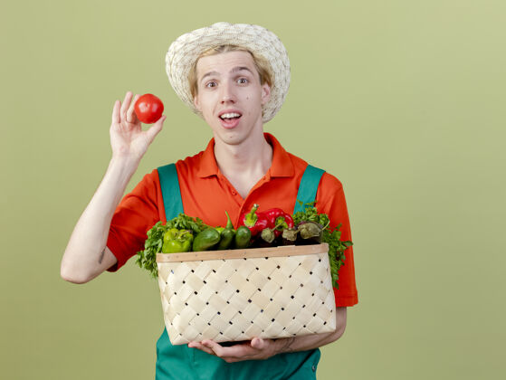 西红柿年轻的园丁穿着连体衣 戴着帽子 手里拿着装满蔬菜的箱子 站在明亮的背景下 西红柿微笑着 脸上洋溢着幸福站着园丁脸