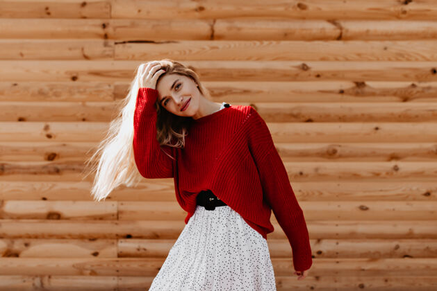 明亮可爱的金发女郎穿着时髦的红色毛衣在外面玩得很开心快乐的年轻女子在木墙上快乐地摆着姿势成人衣服裙子